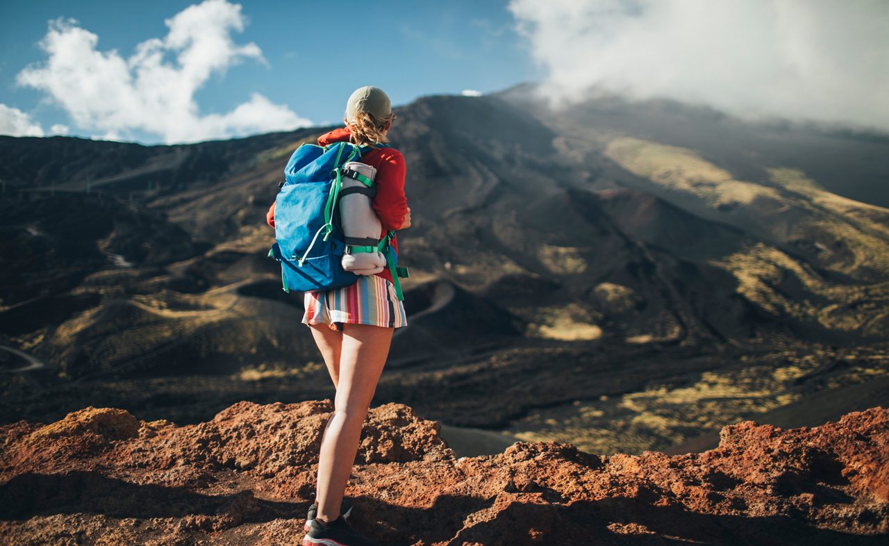 Hiker with a blue backpack standing on a volcanic ridge overlooking Mt. Etna's dark lava fields and slopes under a partly cloudy sky.